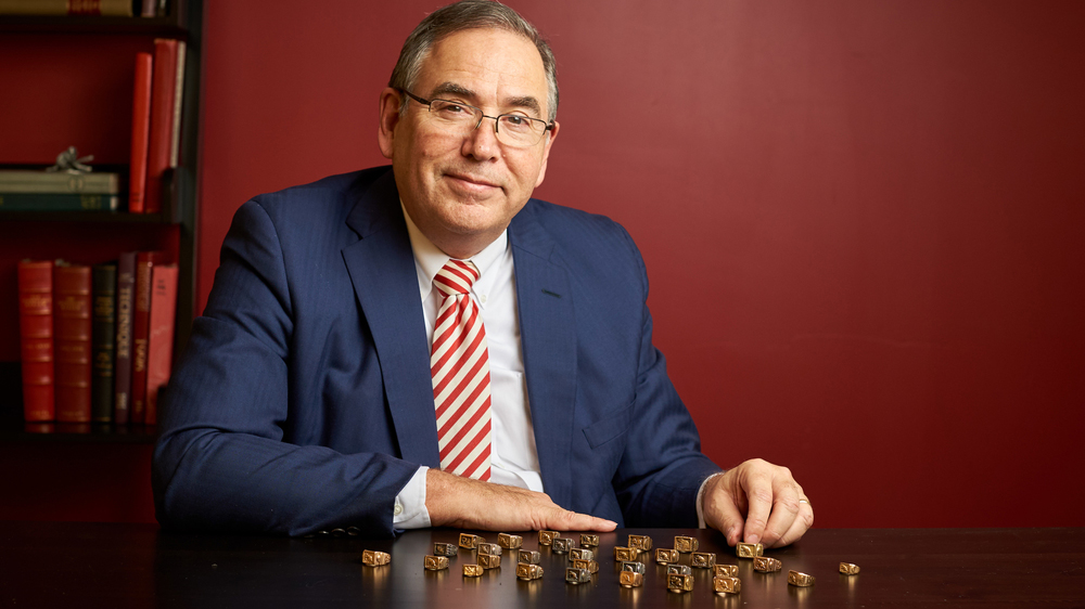 Stephen Fantone wearing a blue suit and sitting at a table in front of a dark red wall