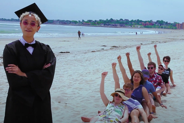 a graduate in cap and gown standing on a beach in front of a row of dancers