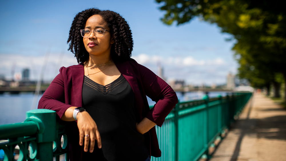 Jordan Harrod standing leaning on a fence next to the Charles River in Cambridge.