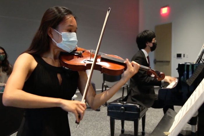 A violinist wearing a mask performs, with a pianist in background