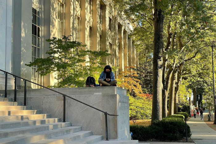 Sunny photo of a person on laptop sitting near front steps of Lobby 7
