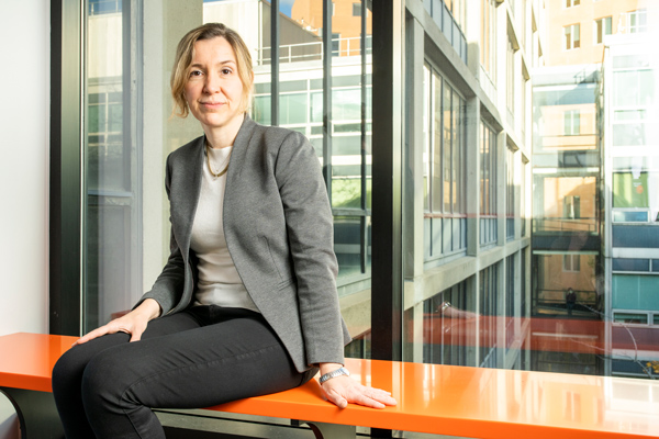 Photo of Asu Ozdaglar sitting on desk with glass walls and Stata in background