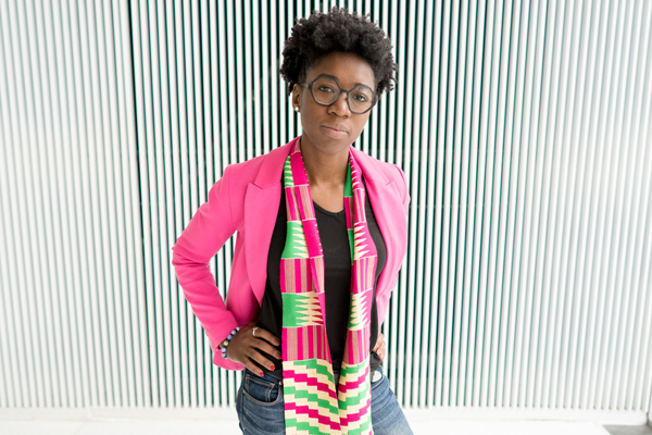 Joy Buolamwini wearing a pink blazer and standing in front of a black and white wall