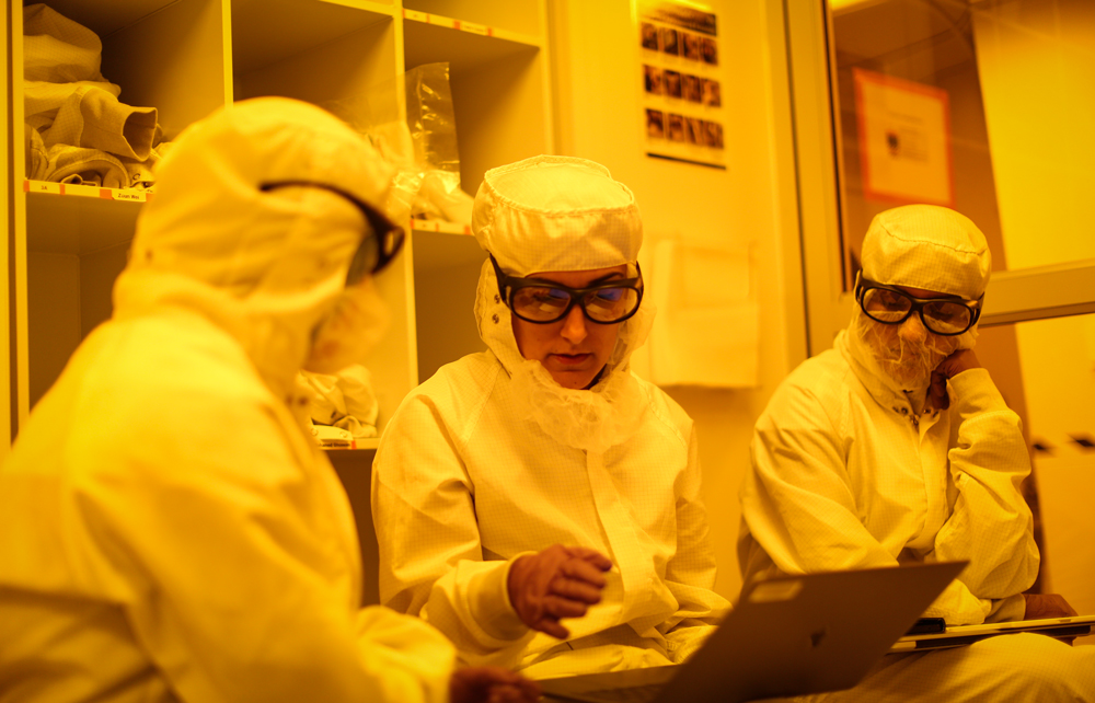 Three people wearing special suits inside a clean lab and bathed in yellow light
