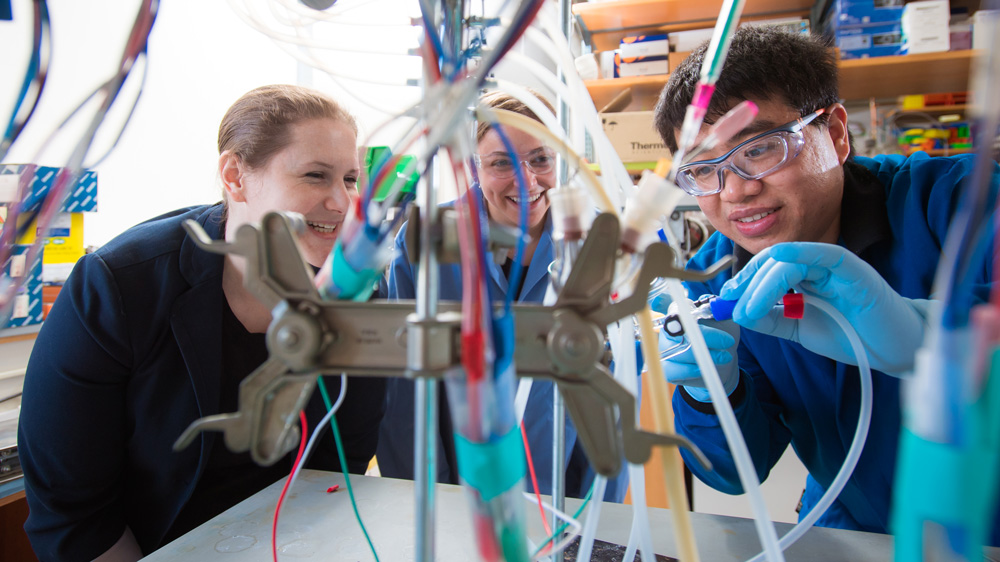 Three smiling people looking at a piece of lab equipment with many tubes
