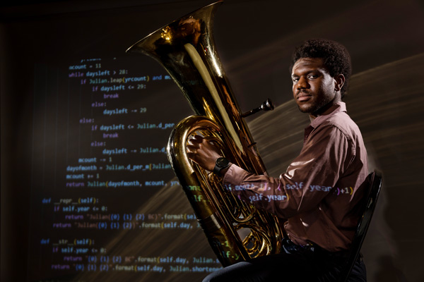 Frederick Ajisafe sits and holds a tuba in a dark room with dramatic lighting. Coding language is projected onto Ajisafe and the wall.