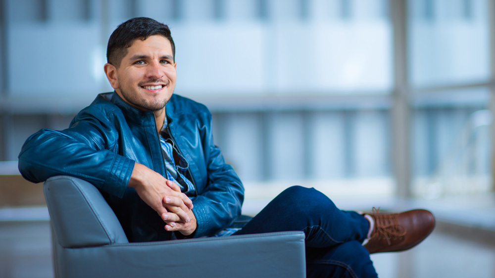 Vila sits on a blue chair with legs crossed, and large windows in background. 