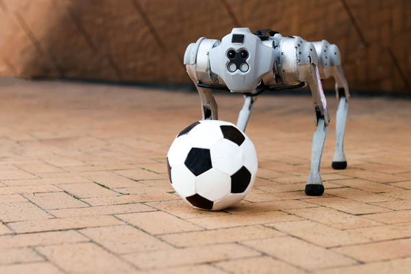 A four-legged, dog-like robot stands next to a soccer ball against an amber background.