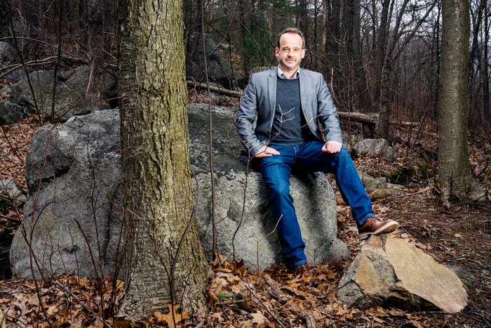 Fournier sits on a large grey rock in the woods. The textures of the trees, rocks, and dead leaves contrasts with Fournier’s blue jeans.