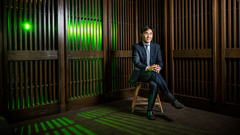 Kim is seated in chair in a room with wooden slatted walls. Through the slats, a green light shines and casts interesting shadows on the ground.