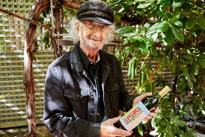 Latif Jiji holds a bottle of wine, while outside in a sunlit yard and near a rhododendron. The wine has a hand painted label showing his brownstone home. 