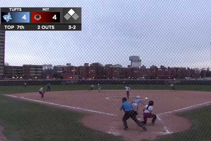 Video still of softball field taken from behind home plate. An MIT softball player slides into home plate. A scoreboard shows the score is "4-4, top 7th, 2 outs, 3-2."