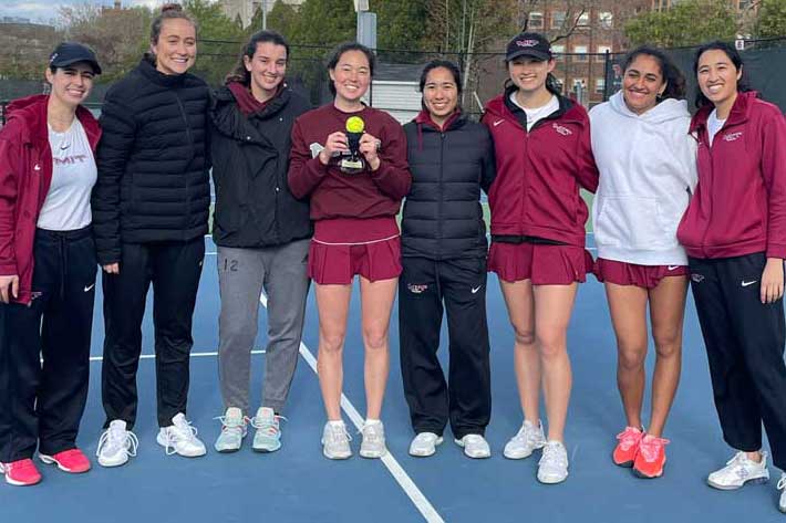 Group photo of 8 members of the MIT Women's Tennis Team on the court, with 1 in center holding a small trohy with a tennis ball inside