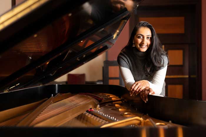 Gurumurthy smiles while leaning on an open grand piano. The inside of the piano is visible, and Gurumurthy’s hands have henna designs.