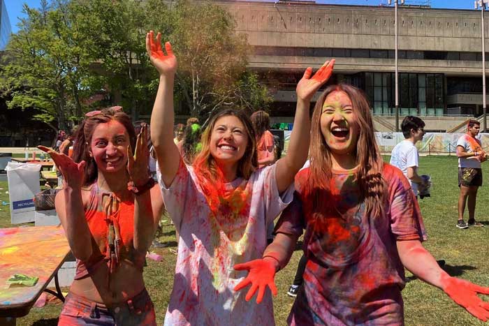 3 people cheer as they throw orange powder in the air, and they are coated with multi-color powders