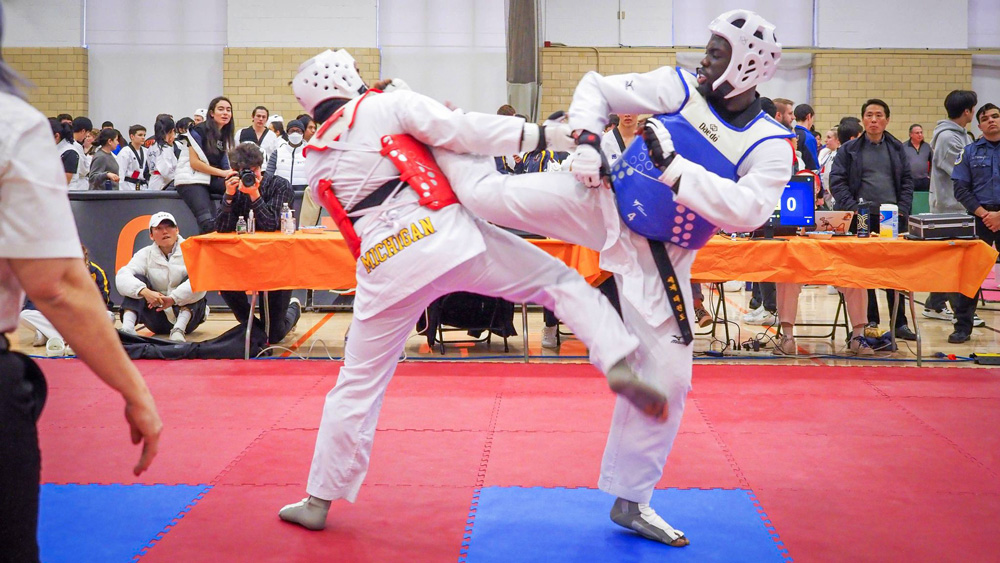 At a competition, Moctar, on right and in blue, kicks dramatically at a competitor in red. The competitor’s uniform says “Michigan.”