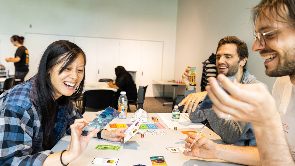 3 people smile excitedly while playing a card game. The game’s cards have blue waves on them, and there is colorful money.