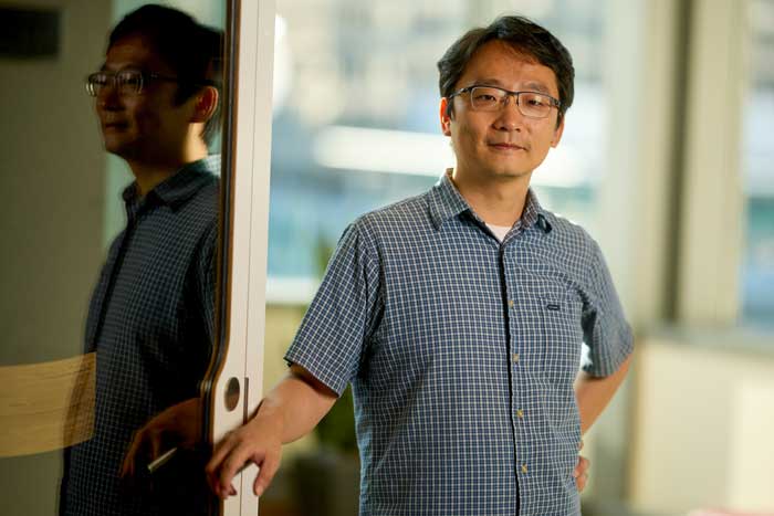 Andy Sun poses for portrait during the daytime next to reflective door while resting their right hand on handle. The blurry background has glass window with view of a building, a red lounge chair, and a round table. 