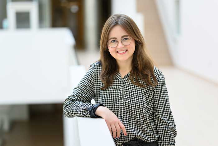 Portrait photo of Kathrin "Kat" Kajderowicz leaning against a railing, with white walls and stair case in background