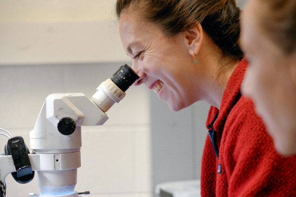 A person looks into a microscope and smiles, as someone else looks on.