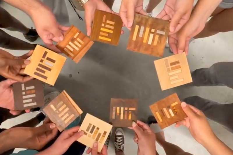 A circle of 10 hands hold wooden tiles that have the MIT logo