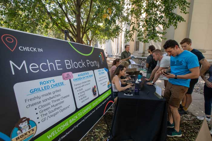 A sign says "MechE Block Party" while students chat near the check-in table