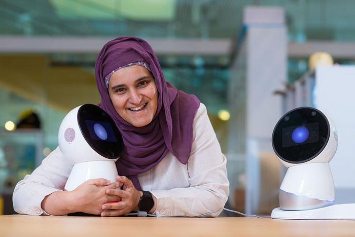 Sharifa Alghowinem, who wears a head scarf, leans on a table, hugging a Jibo personal robot, which looks like a sphere with a large eye.