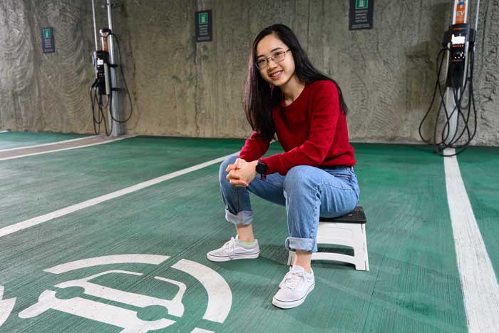 Elaine Siyu Liu sits on a stool inside a parking garage. The ground is painted green and there is an electric car logo. Plus, in the background are electric are charging stations.