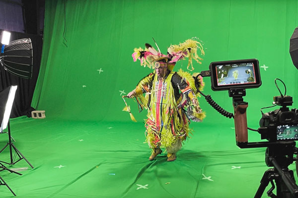 a traditionally dressed Indigenous person dancing in front of a green screen