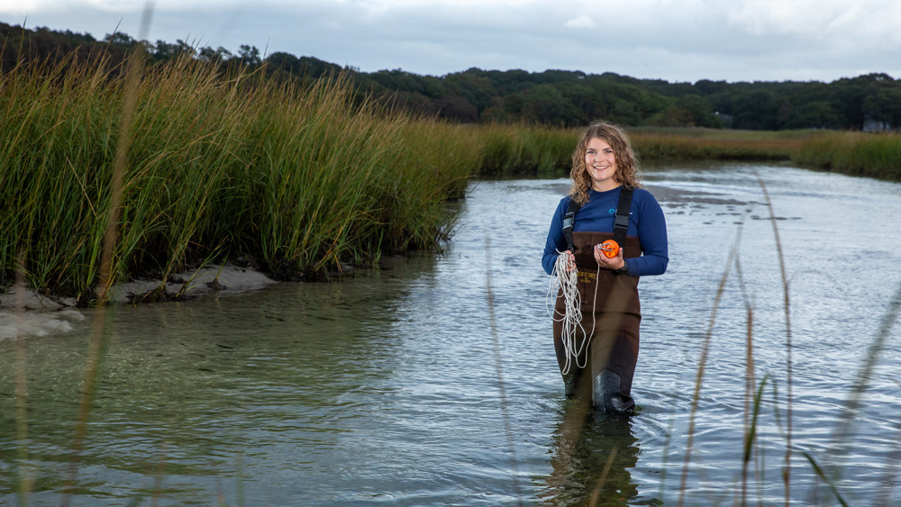 Faith Brooks, wearing waders, stands knee-deep in a pond, holding an instrument and a coil of rope.