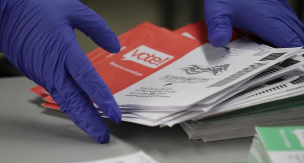 We see a pair oif hands, with purple gloves on, stacking red and white vote by mail ballots.