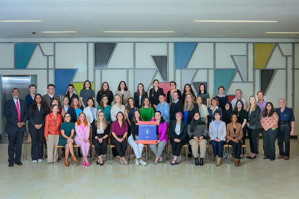 Attendees of the NEA Rising Stars Workshop photographed with organizers, and guest speakers. Two rows standing, front row seated inside in fron of a wall with a multi-colored geometric pattern, MIT