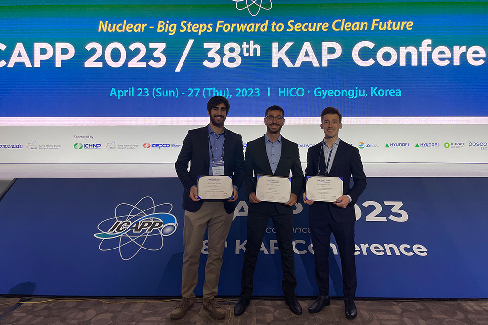 Three male students holding award cetificates standing in front of 2023 ICAPP wall-sized banner, MIT