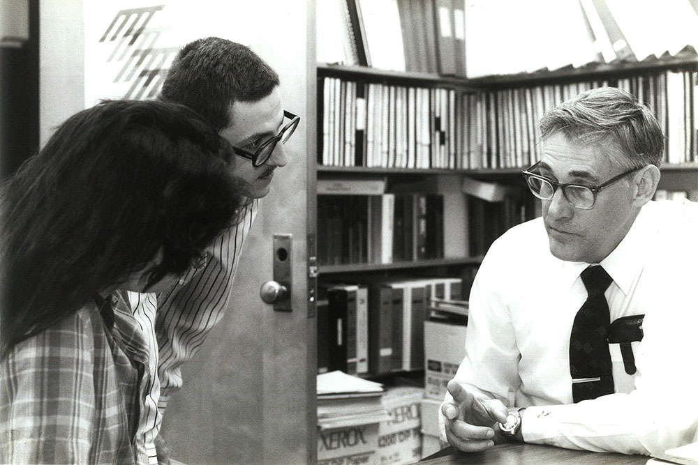 On right, David Lanning seated ;eaning on table in a bookshelf-lined room, one female and one male student on the left in the doorway to the room speaking