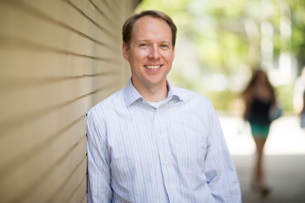 Ben Forget, male faculty member leaning against an outdoor brick wall with blurred greenery in the background, MIT
