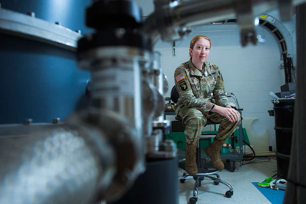 ill Rahon in Army fatigues seated on a swivel chair in a lab with instuments and lab equipment behind her, and our of focus to the front left, MIT