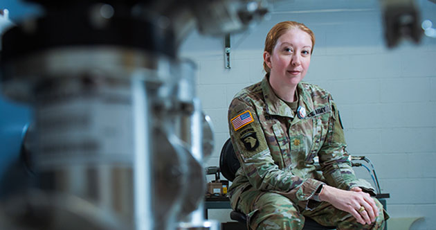 Jill Rahon in Army fatigues seated on a swivel chair in a lab with instuments and lab equipment behind her, and our of focus to the front left, MIT