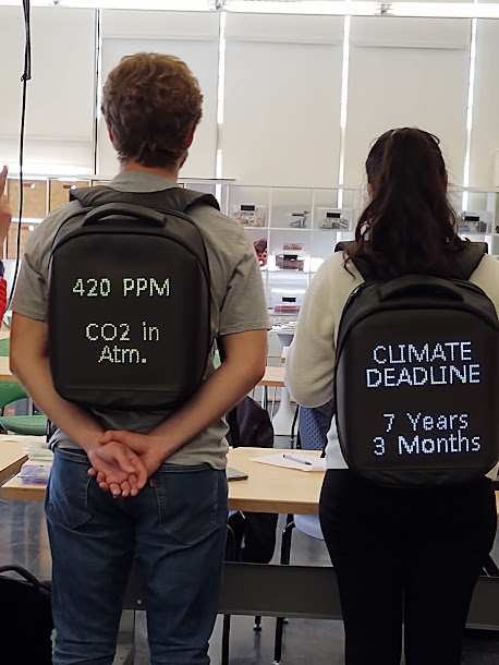 Henri and Jessica with backpacks showing 420ppm CO2 in atm. and climate deadline 7 years 3 months