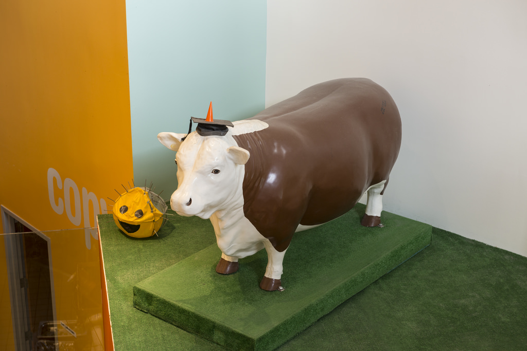 A brown and white life-size cow sits on a green platform in the Stata Center.
