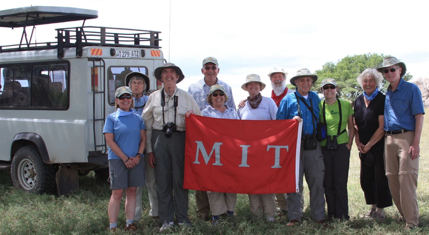 A group of 11 people wearing safari gear stand in front of a van and hold a red MIT banner.