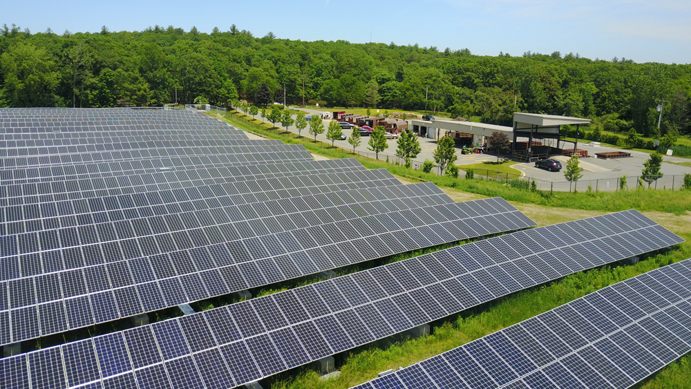 A large solar farm in the middle of a wooded area with a parking lot on the right of the field