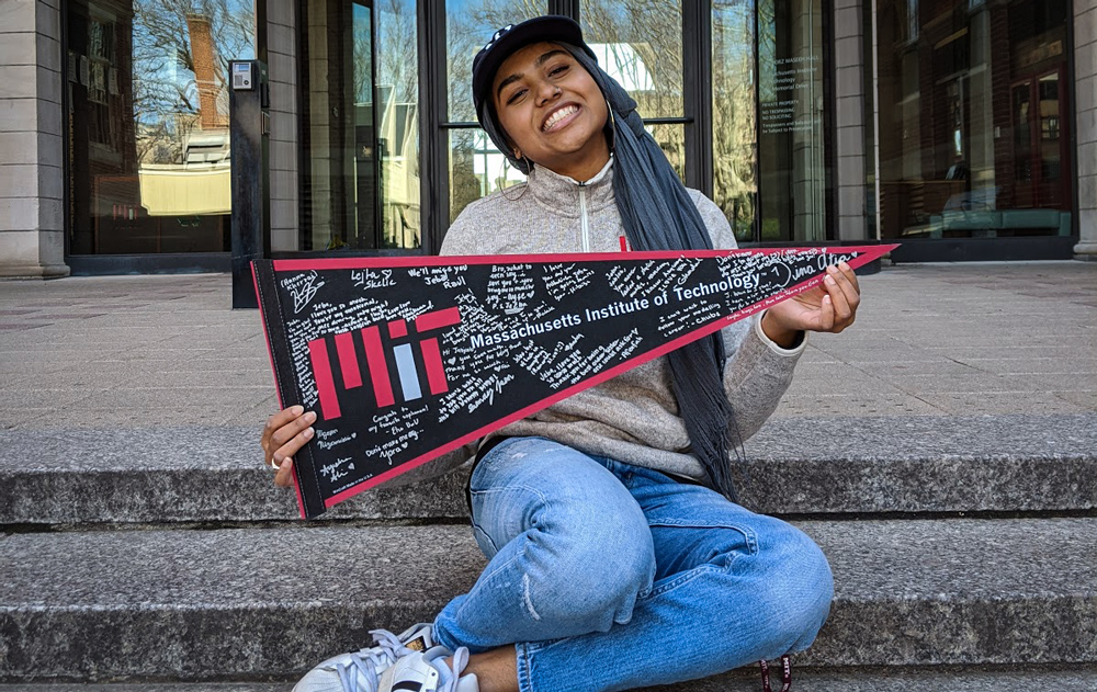 Smiling student at MIT sits on steps of her dorm holding a banner covered in signatures