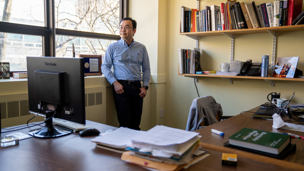 Photo of Yen-Jie Lee near window in his office, with papers, books, and family photos on desk and shelves