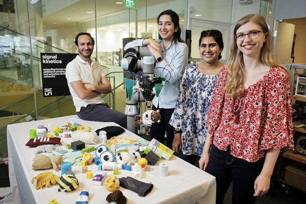 Four researchers standing next to a robotic arm and a table covered with a variety of items