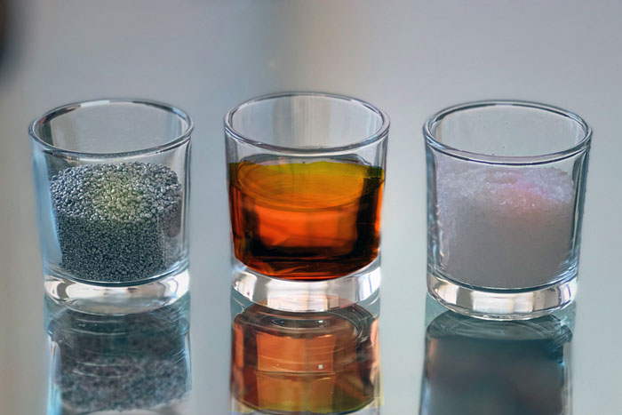 3 small glasses hold 3 different materials: silver pebbles of aluminum (left), liquid brownish sulfur (center), and rock salt crystals (right).