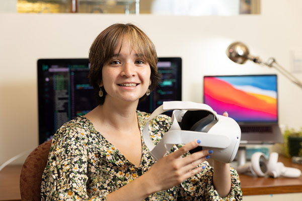 Fernanda De La Torre sits at a desk holding an Oculus Virtual Reality headset, with computers in background.