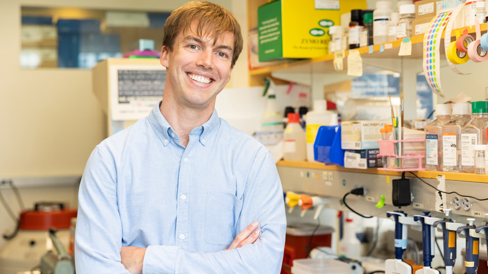 Mitch Murdock smiles in the lab, with shelves and a table filled with colorful equipment and supplies.