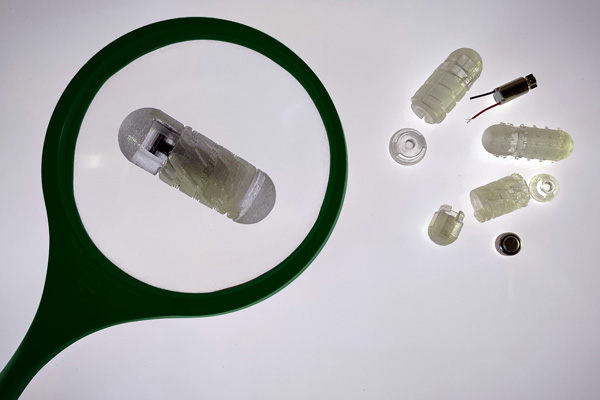 Pieces of the drug capsule prototype are on a grey background. Some pieces are pill-shaped and made of translucent material. Some are threaded like a screw. On the left, a green magnifying glass enlarges one pill-shaped device.