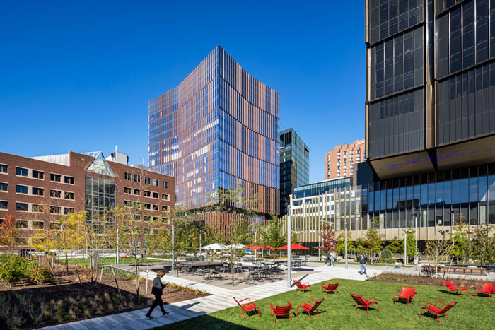 Photo of MIT buildings centered on 314 Main Street, a 17-story glass building that houses the MIT Museum on the first 3 floors. A courtyard with grass, tables, chairs, and walkways is in the foreground.