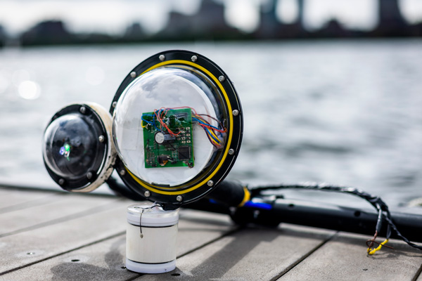 A prototype camera, with 2 bulbous lenses, a circuit board, wires and white cylinder, sits on a dock with the Charles River and Boston skyline in blurred background. 